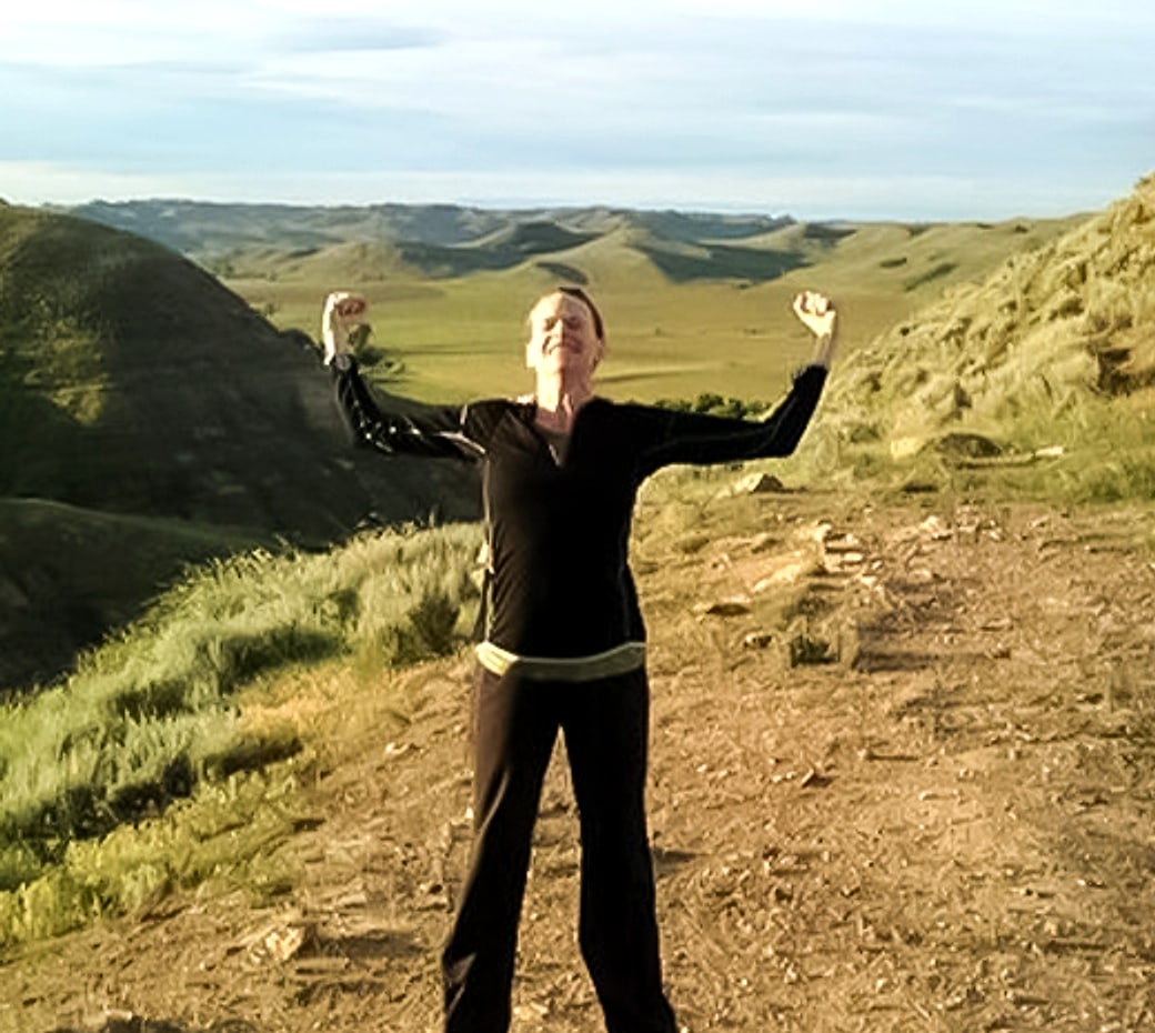 Anna on a grassy ridge with her arms raised over a wide valley